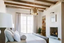 a chic and welcoming Provence bedroom with a built-in fireplace, a grey bed with neutral bedding, wooden nightstands, built-in shelves and wooden beams on the ceiling
