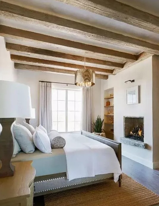 a chic and welcoming Provence bedroom with a built in fireplace, a grey bed with neutral bedding, wooden nightstands, built in shelves and wooden beams on the ceiling