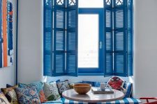 a colorful Mediterranean dining nook with bold blue shutters, a corner sofa with printed pillows, a round table and a chic chandelier