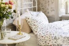 a dreamy Provence bedroom with floral wallpaper, a white metal bed with floral bedding, a white table, a floral bench, artwork and blooms