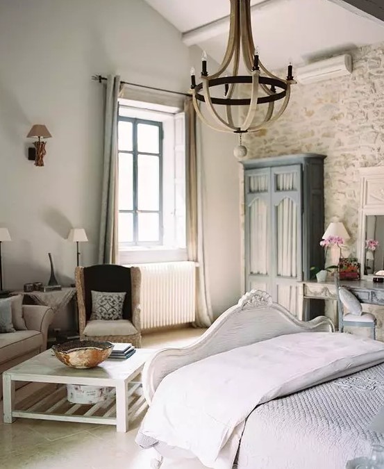 a lovely Provence bedroom with a stone accent wall, a refined white bed, grey seating furniture, a blue wardrobe and a vanity, a wooden chandelier and chic lamps