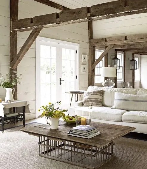 a lovely and welcoming rustic living room done in neutrals, with wooden beams, a wooden table, upholstered furniture and blooms