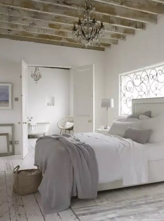 a neutral French country bedroom with wooden beams, a window with a wrought cover, a creamy bed with white and grey bedding, artwork and a crystal chandelier