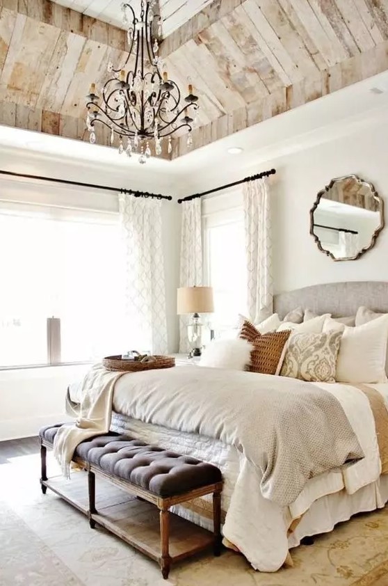 a neutral Provencal bedroom with a wooden ceiling, a grey bed with neutral vintage bedding, an upholstered bench, a crystal chandelier and lots of natural light