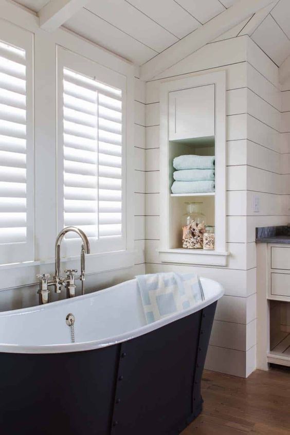 a neutral farmhouse bathroom with planked walls and a ceiling, shutters that match the neutral walls and a navy bathtub