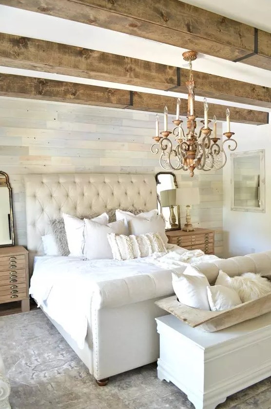 a neutral rustic bedroom with a catchy pastel accent wall, wooden beams on the ceiling, an upholstered neutral bed with neutral bedding, a white storage chest and a chic chandelier