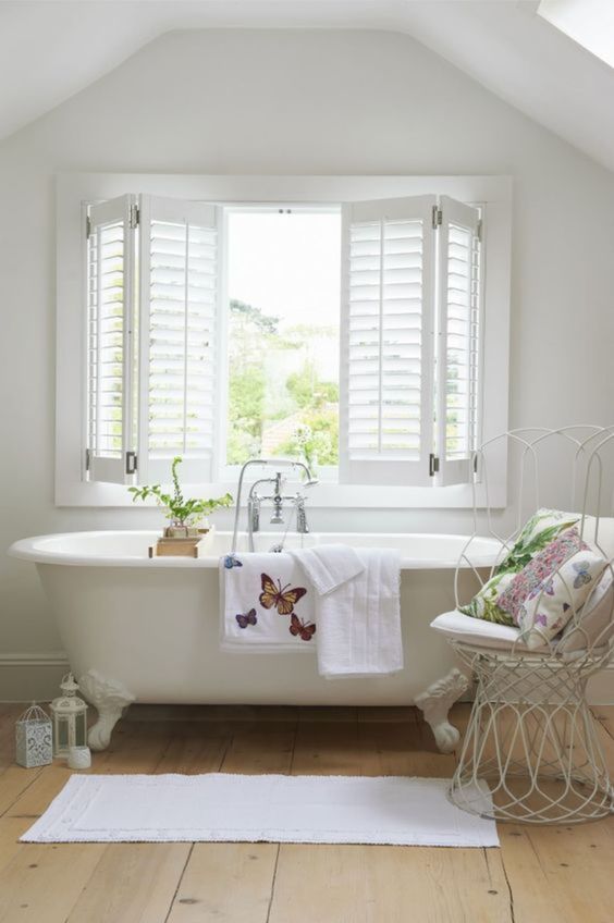a neutral vintage inspired attic bathroom with shutters covering the window for privacy, a creamy clawfoot bathtub, an elegant metal chair