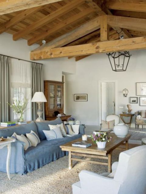 a rustic chic living room with a wooden ceiling and wooden beams, a blue sofa and pillows, a wooden dining table and pastel chairs