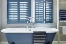a seaside bathroom with all neutral everything, a blue clawfoot bathtub and matching cafe style shutters that echo with it