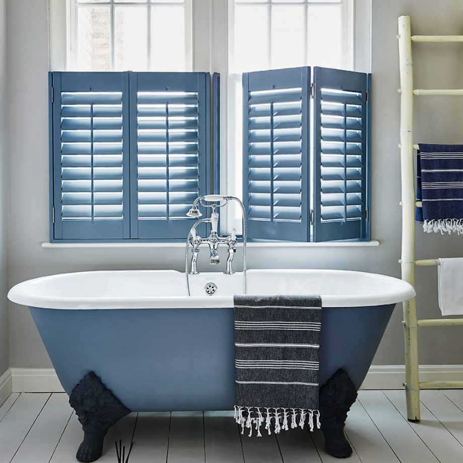 a seaside bathroom with all neutral everything, a blue clawfoot bathtub and matching cafe style shutters that echo with it