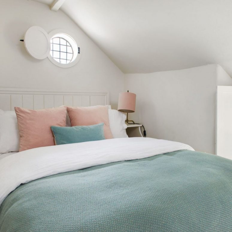 a small neutral attic bedroom with a planked bed and white nightstands, pastel bedding, a round window with a solid shutter