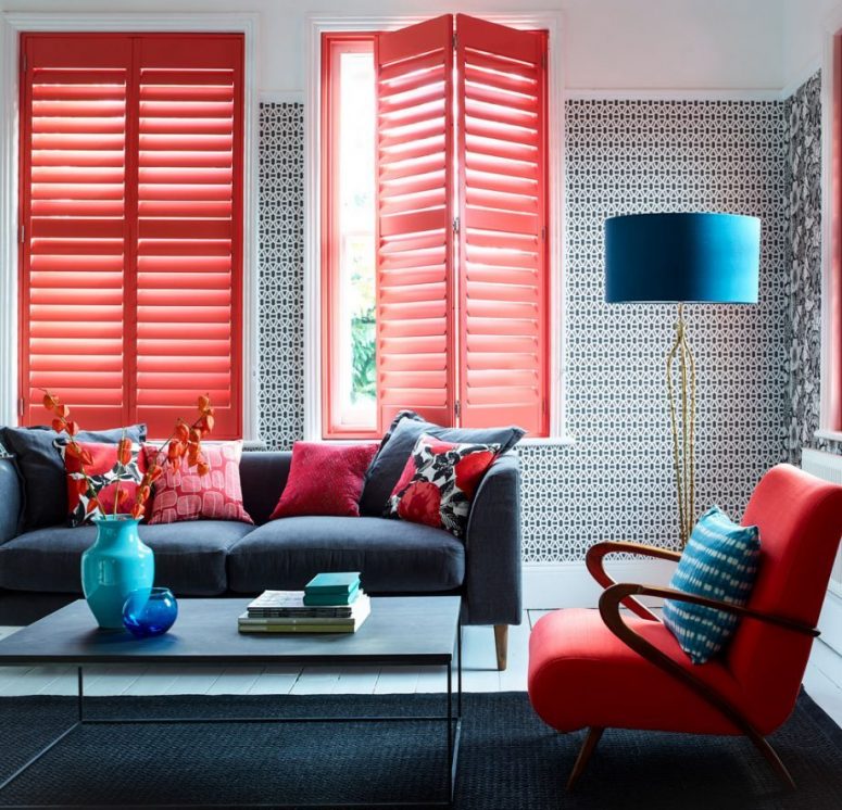 a stylish mid-century modern living room with printed wallpaper, a black rug, a grey sofa with red printed pillows, a red chair and matching red shutters to add extra interest