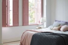a subtle bedroom with planked walls and a floor, a bed with pastel bedding, pink shutters and a nightstand with a table lamp
