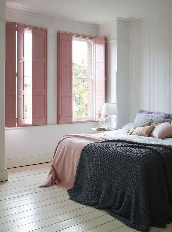 a subtle bedroom with planked walls and a floor, a bed with pastel bedding, pink shutters and a nightstand with a table lamp