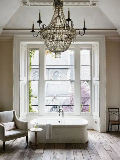 a vintage Provence bathroom with tan walls, a bathtub by the window, a crystal chandelier, a neutral chair and a wooden floor