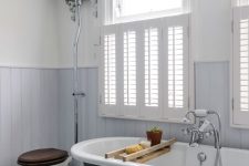 a vintage bathroom with light blue paneled walls, a black clawfoot bathtub, a vintage toilet and shutters covering the part of the window