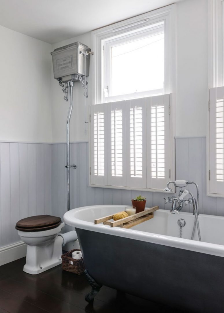 a vintage bathroom with light blue paneled walls, a black clawfoot bathtub, a vintage toilet and shutters covering the part of the window