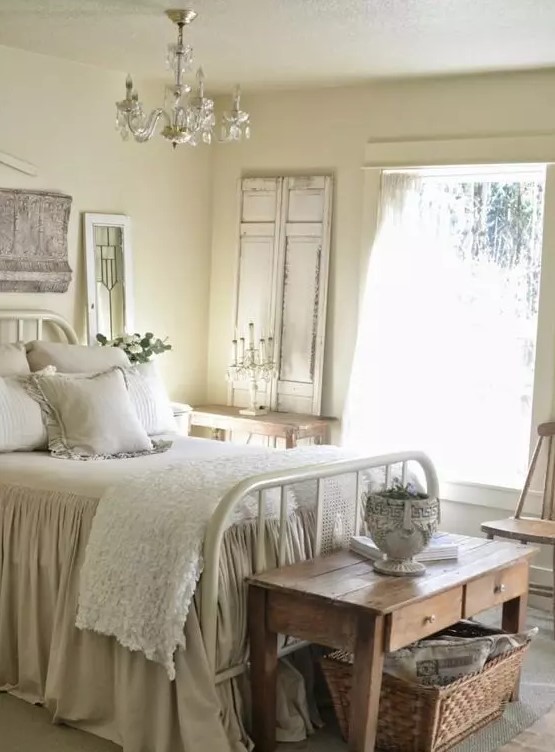 a welcoming Provence bedroom with tan walls, a white metal bed with neutral vintage bedding, a wooden table and a basket for storage, a crystal chandelier