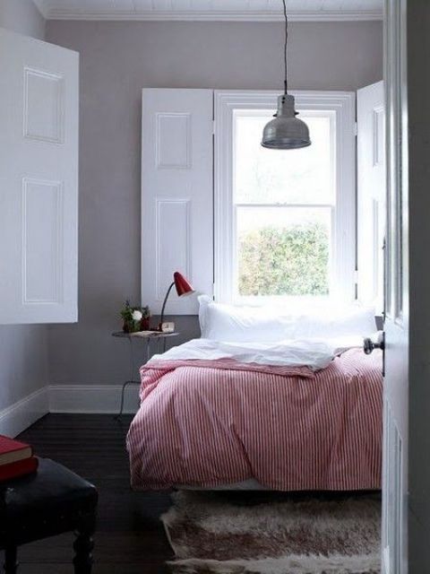 a welcoming bedroom with grey walls, a bed with red and white bedding, a nightstand with a red lamp, a pendant lamp and white solid shutters