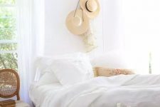 an airy and chic French country bedroom with white walls and a ceiling, a bed with white bedding, a stained chair and a bench, a cool chandelier and straw hats