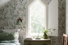 an attic guest bedroom with botanical wallpaper, a bed with green bedding, a house-shaped window with folding shutters and a green dresser