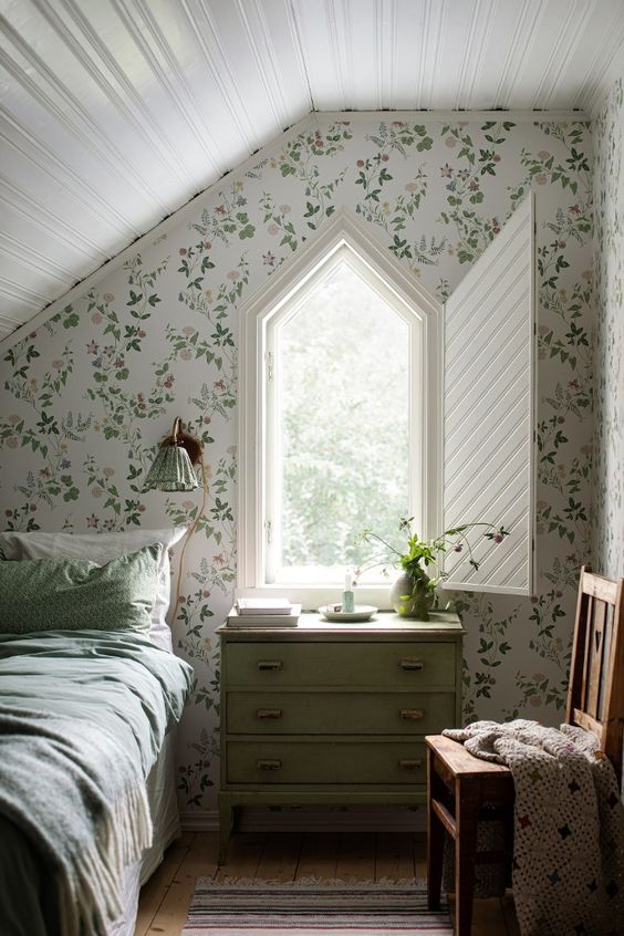 an attic guest bedroom with botanical wallpaper, a bed with green bedding, a house shaped window with folding shutters and a green dresser