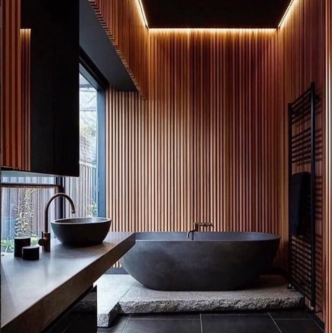 a contemporary bathroom with wood slat walls, a black lit up ceiling, a black bathtub on a stone platform, a black sink and a radiator