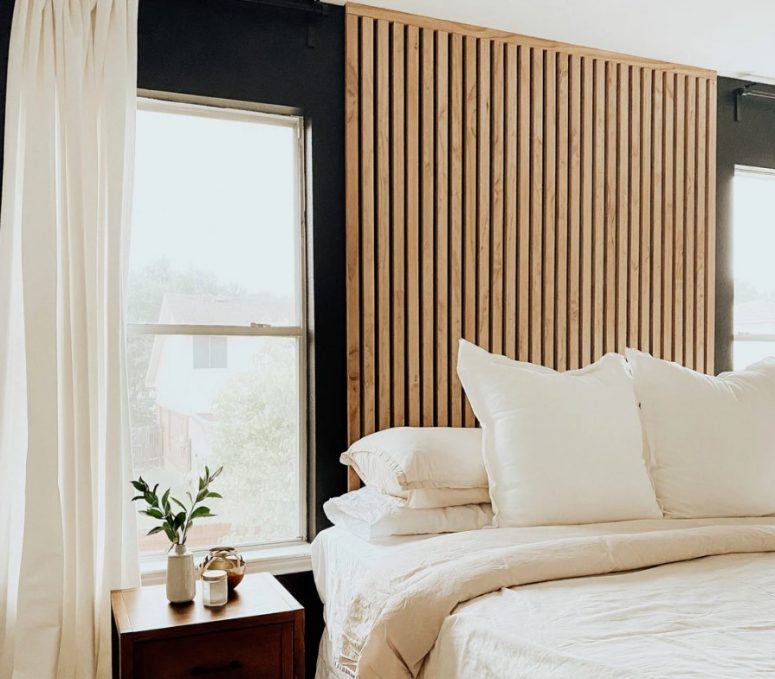 a contemporary bedroom with black walls, a wooden slat accent wall, a bed with neutral bedding, a stained nightstand with greenery and neutral curtains