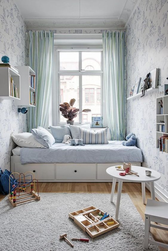 a delicate pastel kid's space with blue wallpaper walls, a neutral bed with blue bedding, a round table, bookshelves and toys