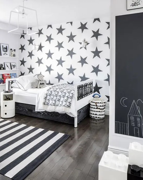 a monochromatic Scandinavian kid's room with a star-printed accent wall, ledges for books, a wihte bed with drawers, a wardrobe with a chalkboard and a striped rug