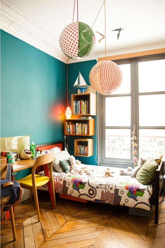 a small and colorful kids' room with a stained bed with printed bedding, emerald walls, bookshelves, a shared desk and colorful chairs