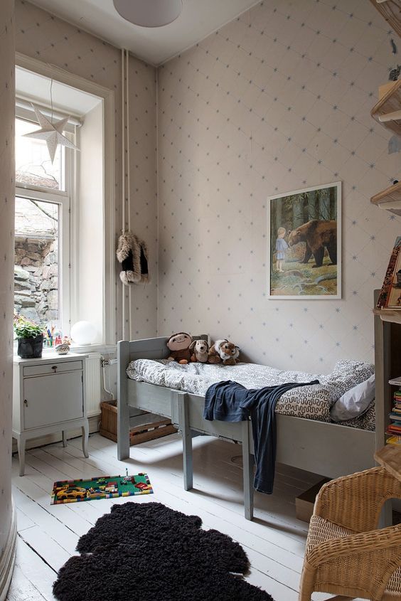 a small Scandi kids' room with printed wallpaper, a grey bed, a white cabinet, open shelves and a wicker chair