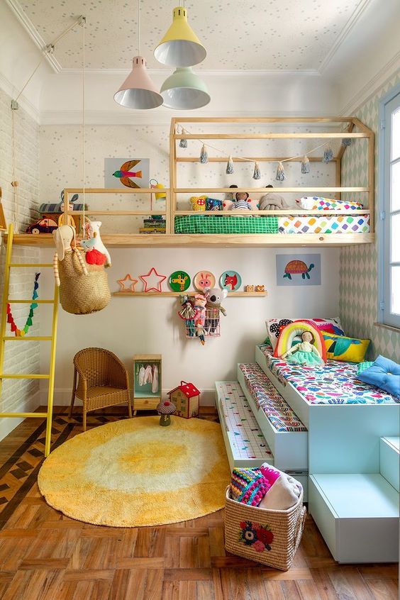 a colorful shared kids' room with a raised bed and a bed at the window, some ladders, a wicker chair and colorful bedding and toys
