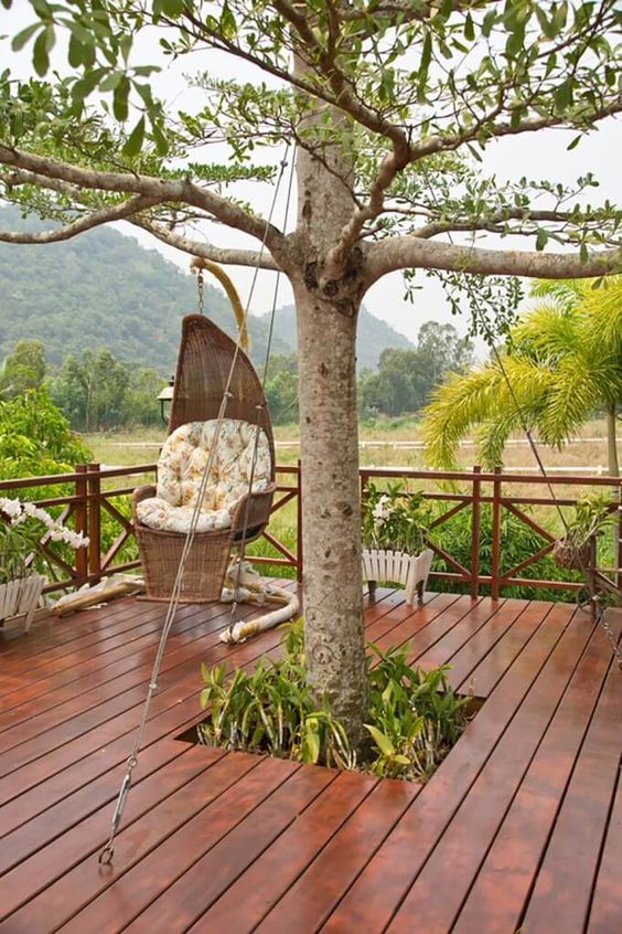 a deck built around a tree, with some greenery surrounding the tree, a wicker pendant chair and some potted blooms