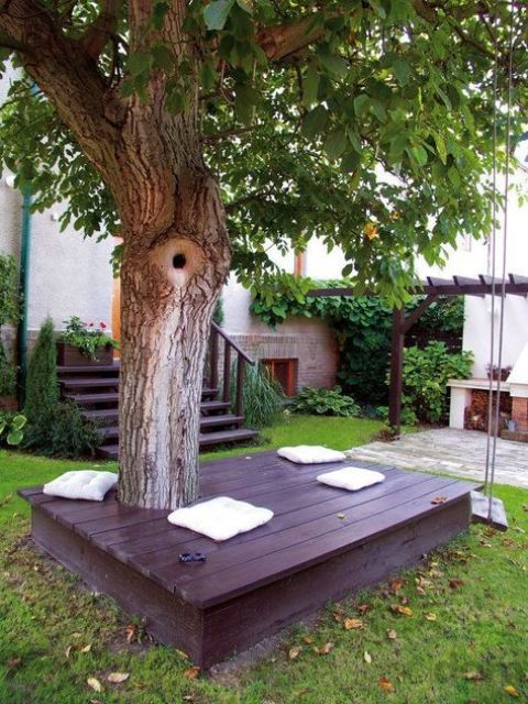a dark deck around the tree with white pillows under the tree is a lovely space that welcomes you to the garden