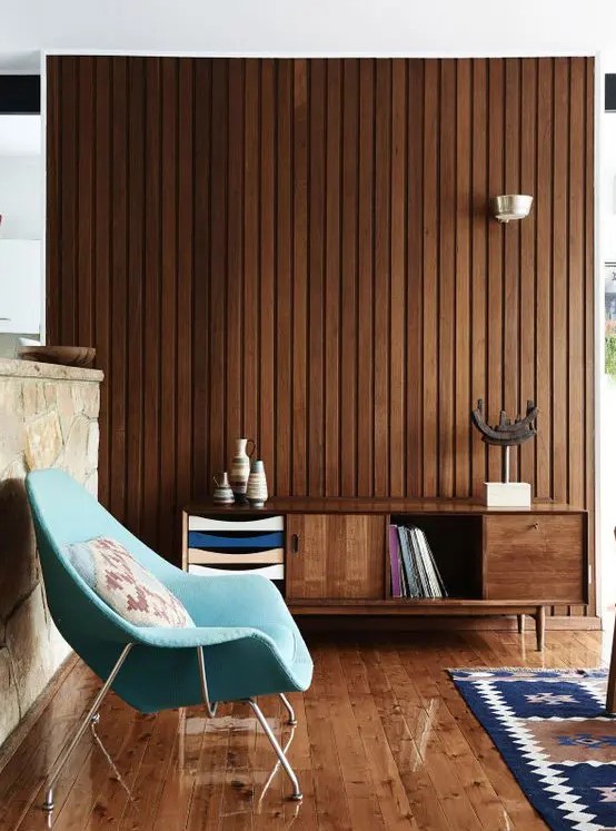 a mid-century modern living room with a rich-stained wooden slat wall, a stained floor and credenza, a blue chair and a printed rug