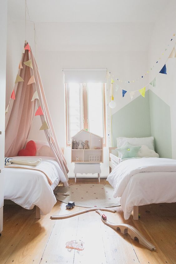 a small shared kids' room with a pink and a green bed and nook, banners, a playhouse, a rug and some simple toys