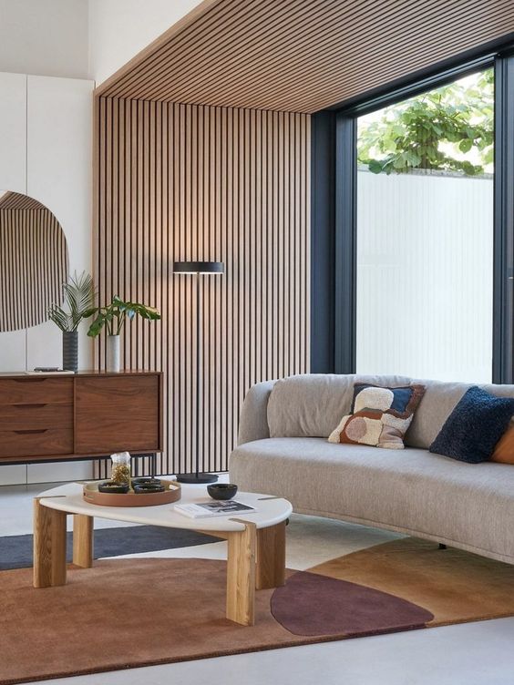 an elegant mid-century modern living room with a wood slat accent wall and ceiling, a curved sofa, a printed rug, a stained credenza and a coffee tabl