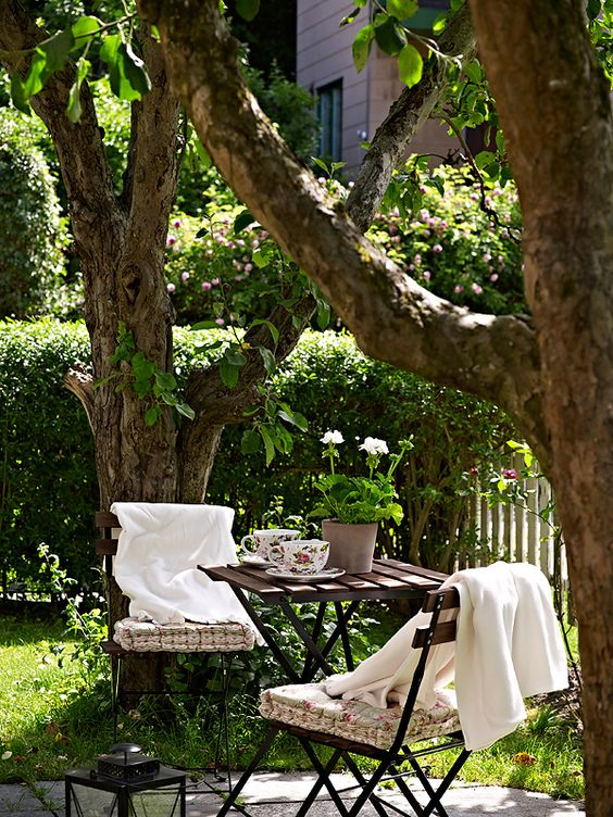 a cozy little dining space around three trees, with a dark stained table, a metal chairs with floral cushions and white blankets