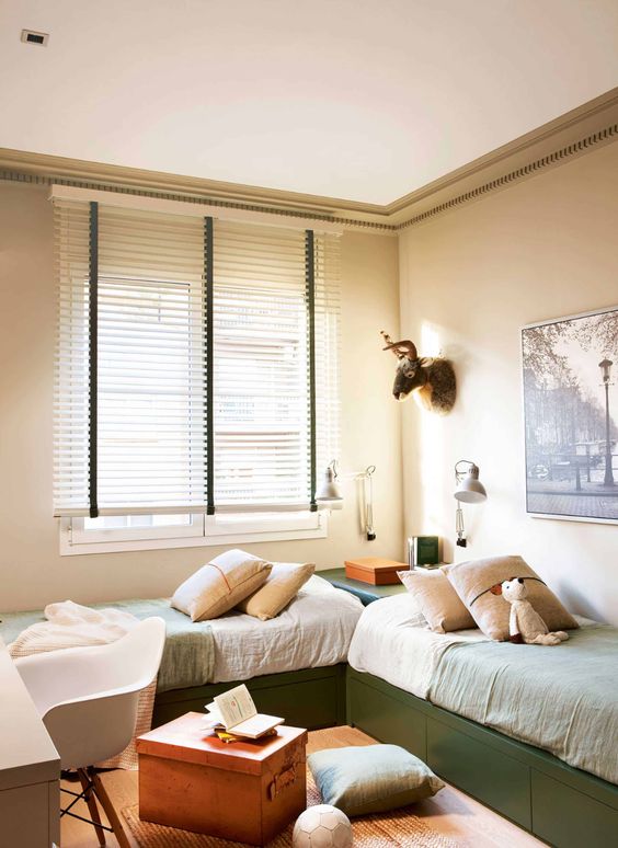 a small shared kids' room with green storage beds, pastel bedding and pillows, a desk and a chair and some artwork