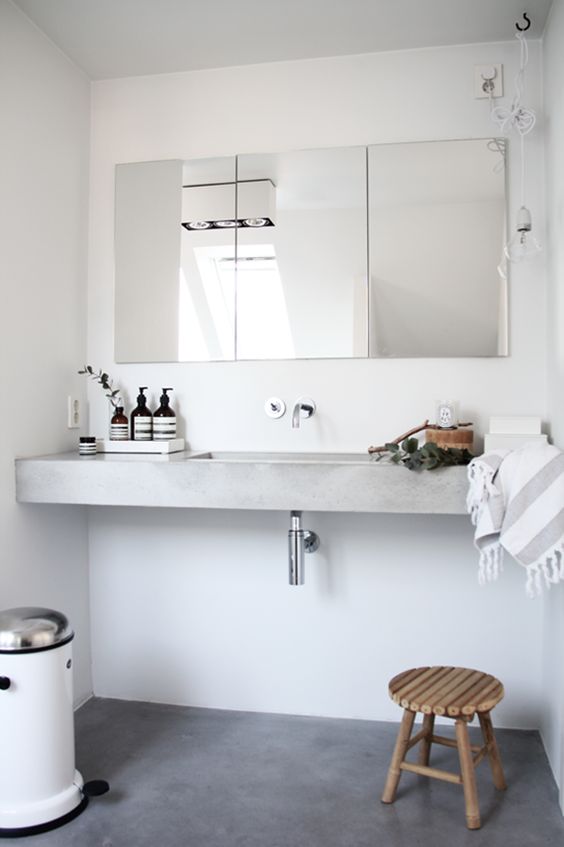 a Scandinavian bathroom done in white, with a floating concrete sink, a mirror cabinet, a wooden stool and a simple metal trash can