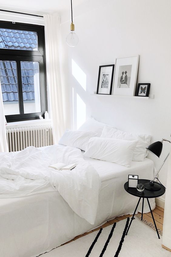 a black and white bedroom with a white bed and white bedding, a black nightstand and a black lamp, a ledge with black and white gallery wall