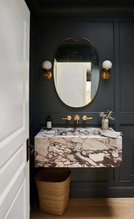 a black bathroom with panels on the wall, a white stone floating sink, brass fixtures and a basket for trash is a lovely and chic space