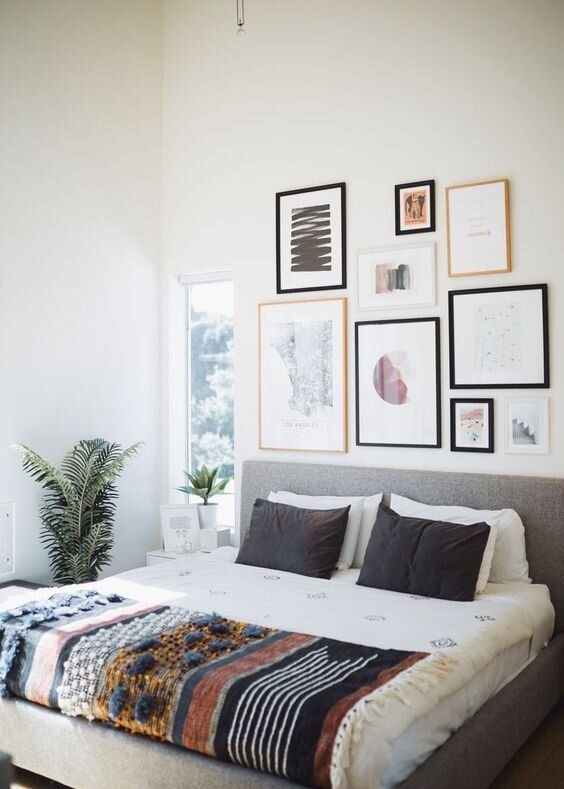 a bright bedroom with a grey upholstered bed and printed bedding, a lovely and fresh gallery wall and some potted plants