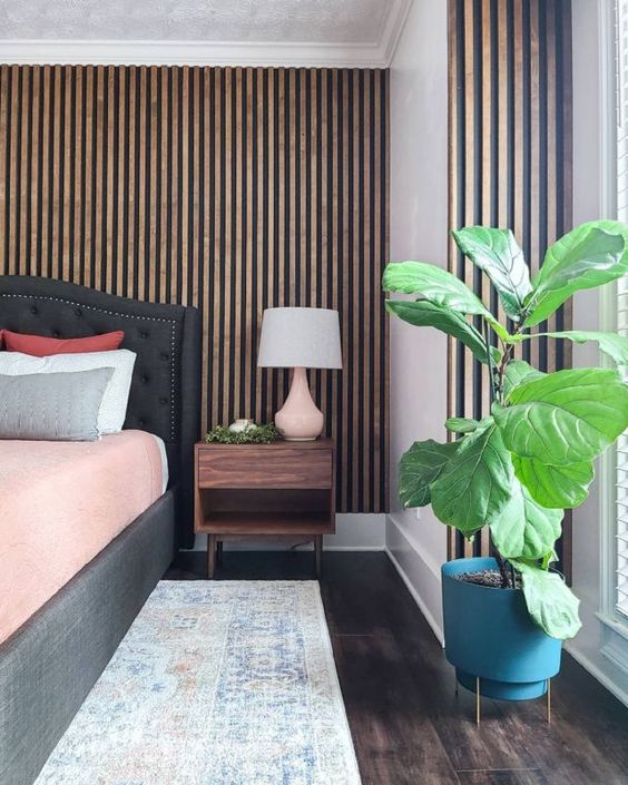 a bright bedroom with a wood slat wall, a graphite grey bed with pink bedding, a boho rug and a potted plant