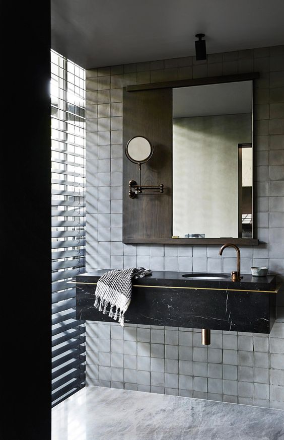 a catchy bathroom done with white square tiles, a black marble floating sink, a mirror with a shelf and some blinds