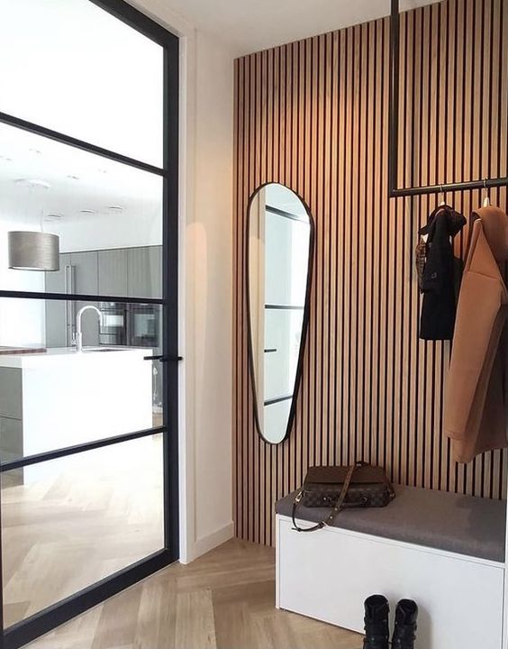 a chic entryway with a wood slat accent wall, a storage bench, a rack and an asymmetrically shaped mirror plus a glass door