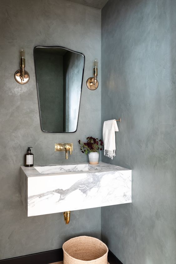 a contemporary bathroom with concrete walls, a floating stone sink, a catchy mirror, gold fixtures and brass sconces is very chic