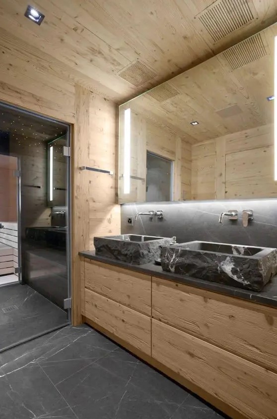 a contemporary chalet bathroom clad with light stained wood, with stone sinks and a countertop, a lit up mirror and a steam room