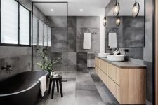 a contemporary grey bathroom clad with concrete tiles, a stone bathtub, a floating vanity, round sinks, black fixtures and framing